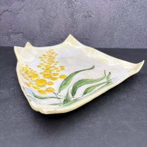 May include: A handmade ceramic serving dish with a floral design. The dish is a light yellow color with a white interior. The design features yellow flower clusters and green leaves. The dish has a unique, asymmetrical shape.