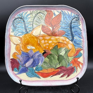 May include: Square ceramic plate featuring a sleeping deer with orange fur and white spots, surrounded by colorful leaves and flowers. The plate has a light pink and purple border and is displayed on a black metal stand. The artwork is hand-painted.