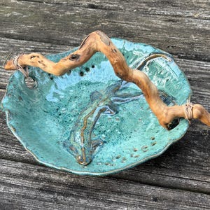May include: A handcrafted, turquoise ceramic bowl with a natural, light brown driftwood accent. The bowl has a textured surface and a unique, organic shape. The driftwood is secured with twine.