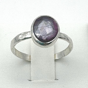 May include: A silver ring featuring a large, faceted, oval-shaped, dark purple gemstone. The ring has a hammered texture on the band, adding a rustic touch. The gemstone is set in a smooth bezel, creating a striking contrast.
