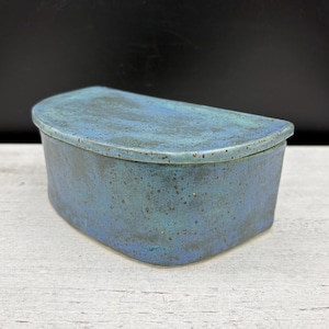 May include: A handmade ceramic box with a lid. The box is a blend of blue and teal hues, with a speckled texture. The lid is curved, and the box has a rectangular shape. The pottery piece is on a white surface.