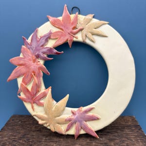 May include: A cream-colored ceramic wreath adorned with colorful maple leaves. The leaves are in shades of red, pink, and tan, arranged around the circular frame. A black hook is attached for hanging. The wreath is displayed on a wooden surface against a blue background.