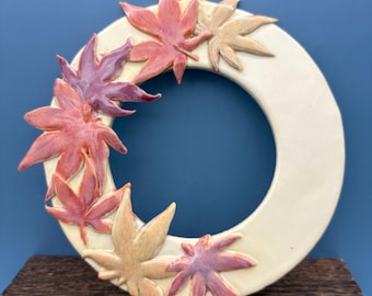 May include: A cream-colored ceramic wreath adorned with colorful maple leaves. The leaves are in shades of red, pink, and tan, arranged around the circular frame. A black hook is attached for hanging. The wreath is displayed on a wooden surface against a blue background.