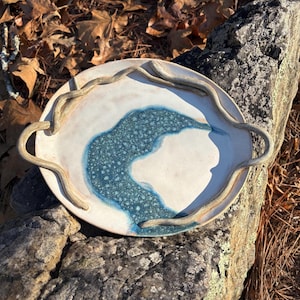 May include: A handmade ceramic serving platter with a white and blue design. The platter has a light beige rim and handles, and a blue, speckled river-like pattern in the center. The platter is resting on a gray rock.