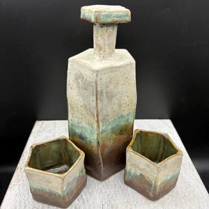 May include: A ceramic decanter with a matching stopper and two matching cups. The set features a hexagonal shape with a reactive glaze transitioning from a dark brown base to a light cream top, accented by streaks of muted green and blue tones.