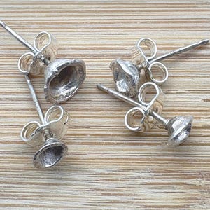 May include: Four silver-colored stud earrings with a bell-shaped design. The earrings are displayed on a light brown wooden surface. The earrings have a textured surface and a post and butterfly backing.