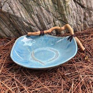 May include: A handcrafted, light blue ceramic bowl with a unique, organic shape. The bowl features a decorative, natural wood handle secured with leather-like ties. The interior has a textured, light-colored ring design. The bowl rests on a bed of pine needles.