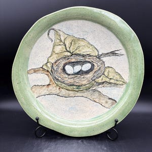 May include: A ceramic plate with a hand-painted bird's nest design. The nest, with three eggs, is nestled on a branch and leaf, set against a speckled white background. The plate has a light green rim and is displayed on a black metal stand.