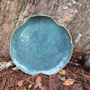 May include: A handcrafted, turquoise ceramic plate with a unique, organic shape. The plate features a textured surface and a wavy rim. The color is a blend of blues and greens, reminiscent of the ocean. Ideal for serving food or as a decorative piece.
