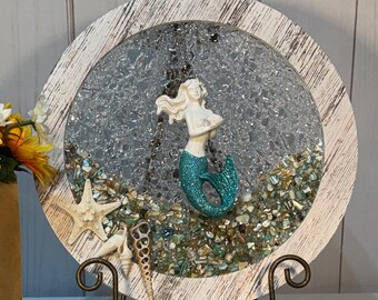 May include: A round, wooden-framed mermaid scene with a white-haired mermaid figure and a teal tail. The scene is filled with clear glass pieces and sits on a decorative metal stand. A brown paper bag with the text "meaghan & mum" holds sunflowers.