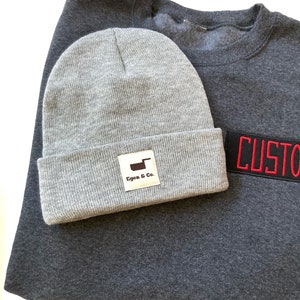K&ouml;nnte beinhalten: Eine graue M&uuml;tze mit umgeschlagenem Rand und einem rechteckigen Etikett mit der Aufschrift "Egon & Co." liegt auf einem dunkelgrauen Sweatshirt. Ein schwarzer Aufn&auml;her mit rotem Text "CUSTOM" ist auf das Sweatshirt gen&auml;ht.