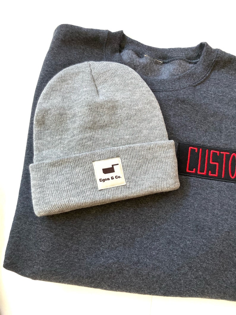 K&ouml;nnte beinhalten: Eine graue M&uuml;tze mit umgeschlagenem Rand und einem rechteckigen Etikett mit der Aufschrift "Egon & Co." liegt auf einem dunkelgrauen Sweatshirt. Ein schwarzer Aufn&auml;her mit rotem Text "CUSTOM" ist auf das Sweatshirt gen&auml;ht.