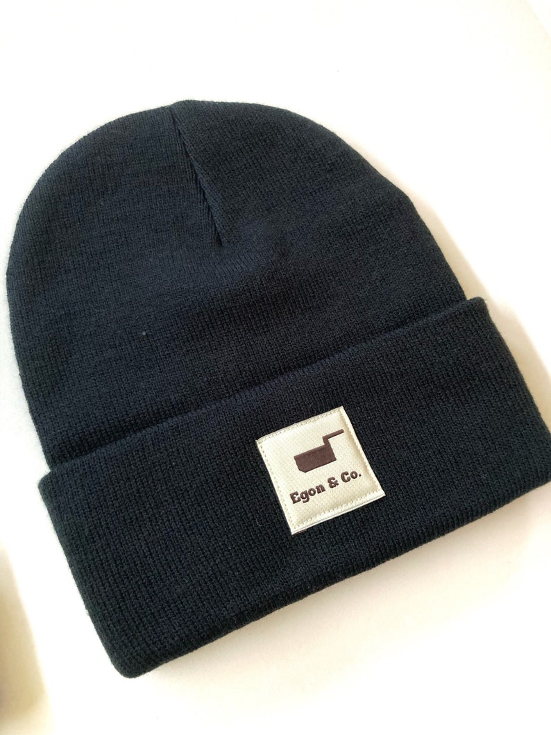 K&ouml;nnte beinhalten: Eine marineblaue Strickm&uuml;tze mit umgeschlagenem Saum. Ein quadratischer, hellbeiger Aufn&auml;her ist auf dem Saum aufgen&auml;ht und zeigt den Text "Egon & Co." und eine braune Grafik. Die M&uuml;tze ist aus einem weichen Strickmaterial gefertigt.