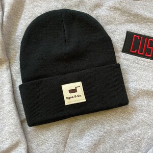 K&ouml;nnte beinhalten: Eine schwarze Beanie-M&uuml;tze mit umgeschlagenem Saum. Ein wei&szlig;es quadratisches Etikett mit dem Text "Egon & Co." und einer braunen Grafik ist vorne aufgen&auml;ht. Die M&uuml;tze liegt auf einem grauen Sweatshirt.