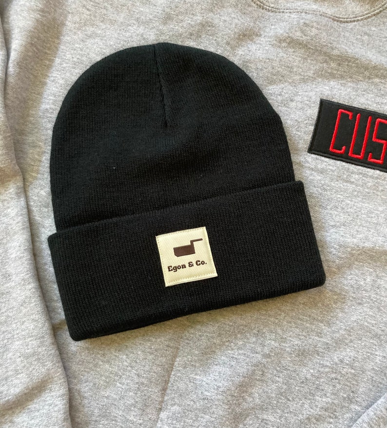 K&ouml;nnte beinhalten: Eine schwarze Beanie-M&uuml;tze mit umgeschlagenem Saum. Ein wei&szlig;es quadratisches Etikett mit dem Text "Egon & Co." und einer braunen Grafik ist vorne aufgen&auml;ht. Die M&uuml;tze liegt auf einem grauen Sweatshirt.