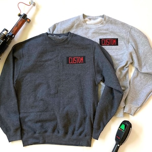 May include: Two sweatshirts with the word "CUSTOM" embroidered on the front. The sweatshirt on the left is dark gray and the sweatshirt on the right is light gray.