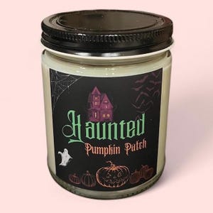 May include: A glass jar candle with a black lid and a label featuring a purple haunted house, a white ghost, orange pumpkins, and bats. The text on the label reads Haunted Pumpkin Patch in green and orange lettering against a black background.