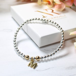Könnte beinhalten: Ein silbernes und goldenes Perlenarmband mit einem goldenen Buchstaben "M" Anhänger. Das Armband liegt auf einer weißen Holzoberfläche.
