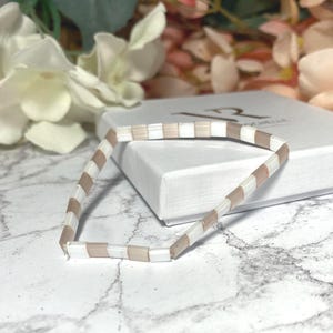 Könnte beinhalten: Ein Armband mit abwechselnd rechteckigen Perlen in Weiß und Hellbraun. Das Armband liegt auf einer weißen Schachtel. Der Hintergrund umfasst unscharfe Blumen. Auf der Schachtel steht das Wort "CHELLE".