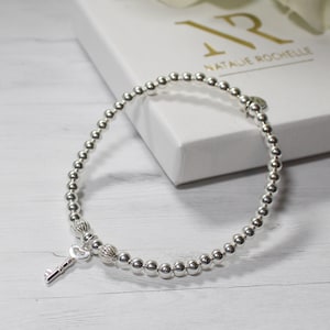 Può includere: Un braccialetto in argento con perline, un ciondolo a forma di chiave e un ciondolo a forma di cuore. Il braccialetto è su una superficie bianca con una scatola bianca su cui è scritto "Natalie Rochelle".