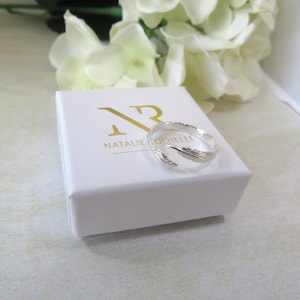 以下が含まれることがあります： 白い箱の上に、羽のデザインのシルバーリングが置かれています。箱には「NR NATALIE ROCHELLE」と金色の文字が書かれています。リングには、2つの詳細な羽のアクセントがあります。背景には白い花があります。