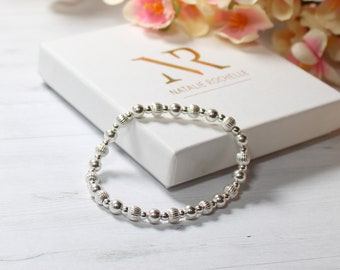 Könnte beinhalten: Ein silbernes Armband mit Perlen, die ein strukturiertes Design aufweisen. Das Armband liegt auf einer weißen Schachtel mit dem aufgedruckten Text "Natalie Rochelle".