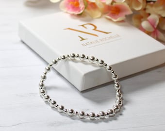 Puede incluir: Un brazalete de cuentas de plata con un diseño sencillo. El brazalete está sobre una superficie blanca con una caja blanca que dice "Natalie Rochelle" en letras doradas.