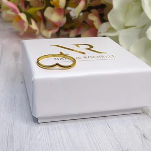 Peut inclure: Une bague dorée en forme de V, posée sur une boîte blanche avec des lettres dorées. La boîte porte l'inscription "NATALIE ROCHELLE". La bague est mise en valeur sur un fond de fleurs roses et blanches.