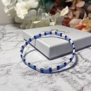 Puede incluir: Una pulsera delicada con un patrón repetitivo de cuentas rectangulares blancas, azul claro y azul oscuro. La pulsera descansa sobre una caja blanca con el nombre de la marca "NATALIE" visible. El fondo presenta flores borrosas y una superficie de mármol.