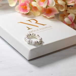 May include: A white box with the text "NR NATALIE ROCHELLE" in gold lettering. A silver ring with sparkly beads is on top of the box.
