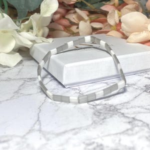 Op de afbeelding: Een delicate armband met afwisselend grijze en witte rechthoekige kralen. De armband wordt gepresenteerd op een witte doos, met een achtergrond met marmerpatroon. Bloemenaccenten voegen een zachte toets toe.