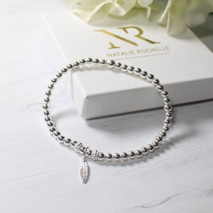 May include: A silver beaded bracelet with a small feather charm. The bracelet is on a white surface with a white box that says "Natalie Rochelle" in gold lettering.