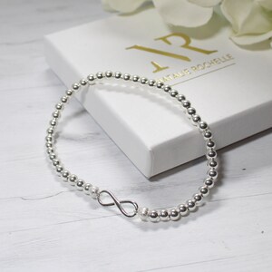 May include: A silver beaded bracelet featuring an infinity symbol. The bracelet is composed of small, spherical, polished silver beads. The infinity charm is a minimalist design. The bracelet is presented on a white box with gold lettering that reads "NATALIE ROCHELLE".