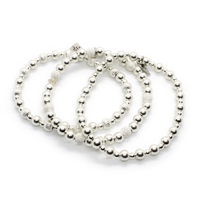 May include: Three silver beaded bracelets with different textures. The bracelets are stacked on top of each other.