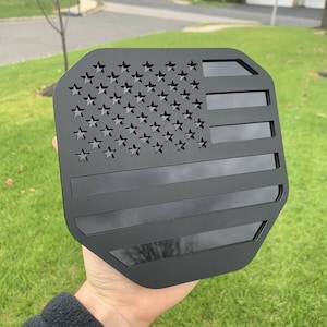 Könnte beinhalten: Schwarzer Metallausschnitt einer amerikanischen Flagge in sechseckiger Form. Die Flagge besteht aus einem massiven schwarzen Material mit einem ausgeschnittenen Design aus Sternen und Streifen.