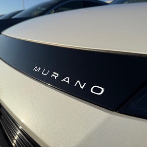 May include: Close-up of a white car hood with a black accent. The word "MURANO" is displayed in silver lettering across the black section. The car is parked outside on a sunny day.