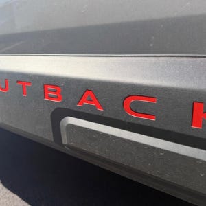 May include: Close-up of a gray vehicle's side panel with the word "OUTBACK" in bright red, bold lettering. The letters are raised and stand out against the textured surface. The image is taken in daylight.