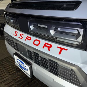 May include: A gray car with a black grille and the word "SPORT" in red letters on the front bumper.