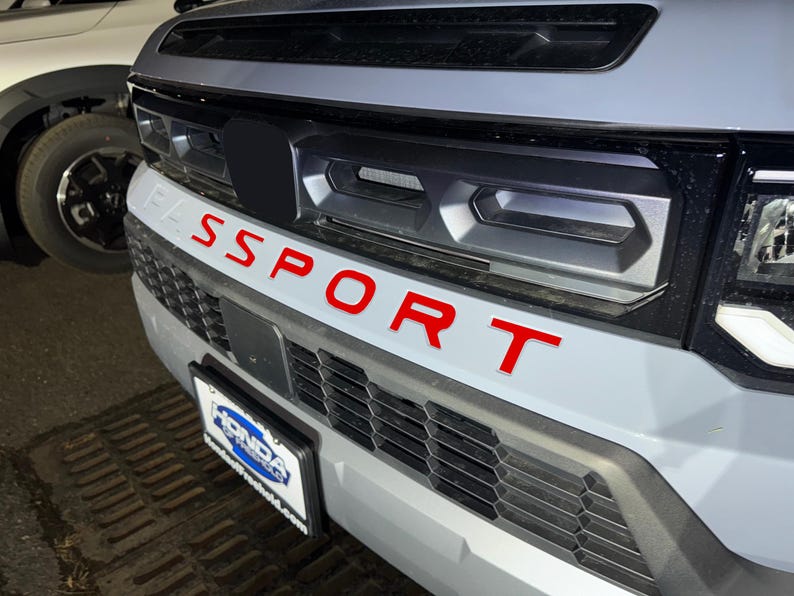 May include: A gray car with a black grille and the word "SPORT" in red letters on the front bumper.
