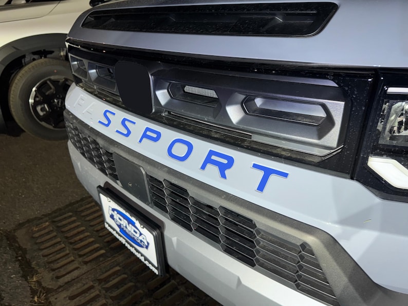 May include: A gray car with a black grill and a blue "SPORT" emblem on the front bumper. The car has a black license plate frame with the word "HONDA" on it.