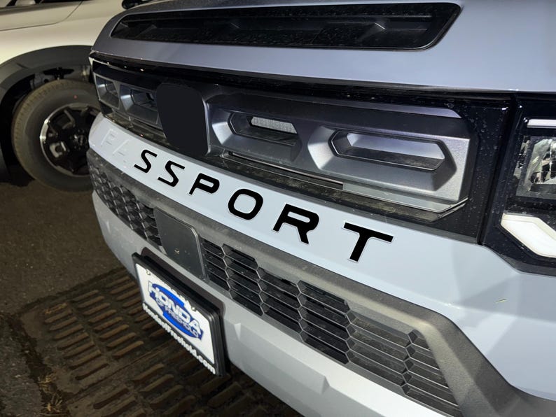 May include: A gray car with a black grille and the word "SPORT" written in black letters on the front bumper. The car has a black license plate frame with the word "HONDA" on it.