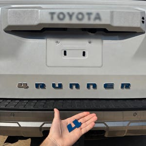 May include: A blue plastic number 4 emblem held in front of the rear tailgate of a white Toyota 4Runner vehicle. The vehicle features the word TOYOTA embossed on the upper trim and the word 4RUNNER in blue lettering across the centre panel.