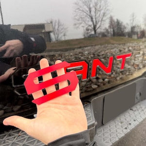 May include: A bright red, stylized letter "S" emblem is held in front of a black vehicle. The vehicle's tailgate features the word "Raptor" in red lettering. The emblem is a custom car accessory.