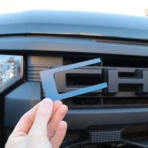 May include: A blue plastic template for a car grille emblem. The template is shaped like a letter C and is being held in front of a black truck grille with the word "CHEV" visible.