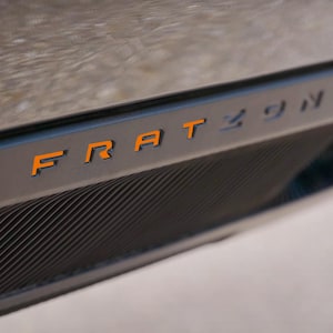 May include: A close-up shot of a dark grey car part, featuring the word "FRATZONIS" in orange. The component has a textured, ribbed pattern and a modern design. The background is out of focus.