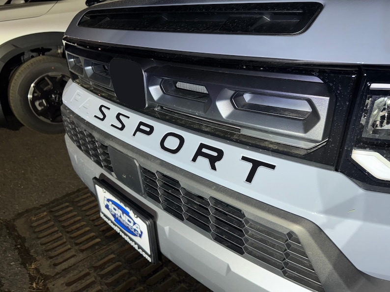 May include: A gray car with a black grille and the word "SPORT" written in black letters across the front. The car has a black license plate frame with the word "HONDA" printed on it.