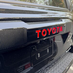 Pode incluir: Close-up da traseira de um veículo preto com o emblema "TOYOTA" vermelho. O veículo tem um suporte de placa preto e um para-choques texturizado. A imagem é tirada ao ar livre.
