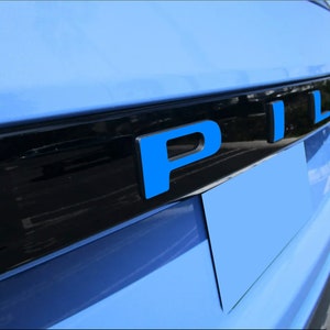 May include: Close-up of a light blue car with the word "PIL" in bright blue letters. The letters are mounted on a black trim, with a light blue background. The image is taken from a low angle.