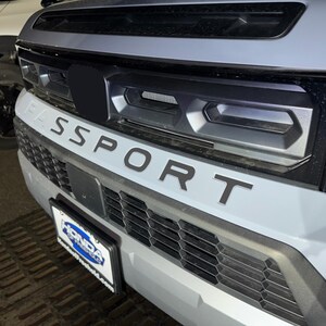 May include: A close-up of the front of a gray SUV with a black grille and the word "SPORT" in black lettering on the front bumper.