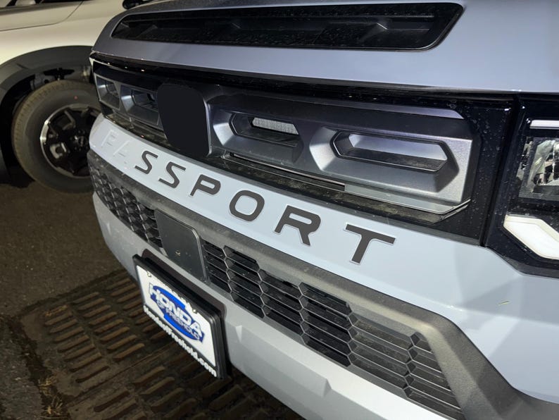 May include: A close-up of the front of a gray SUV with a black grille and the word "SPORT" in black lettering on the front bumper.